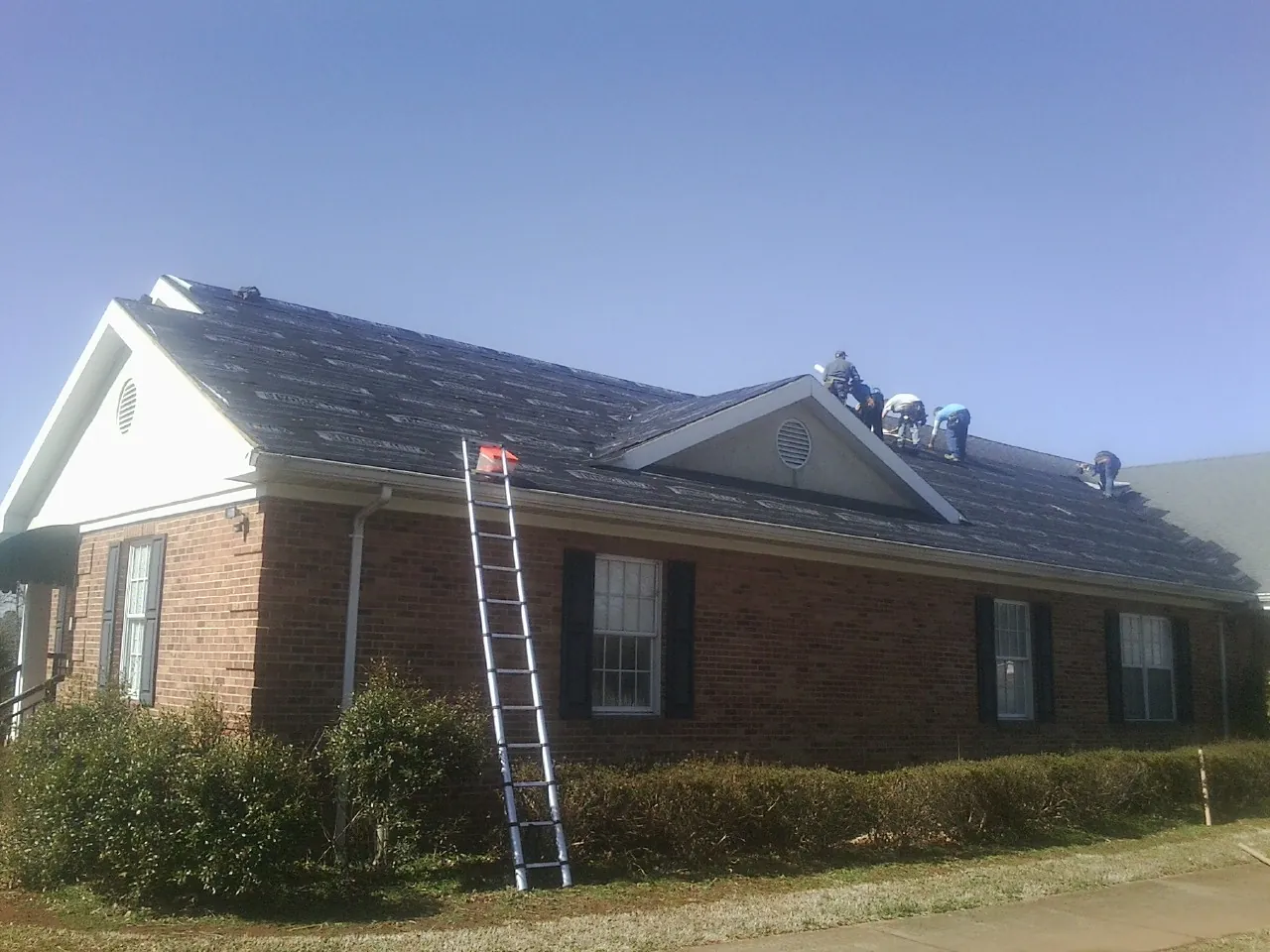 Crew tearing shingles off a brick ranch roof with a ladder set — Metal Roof Repair work by our crew in Blanchard