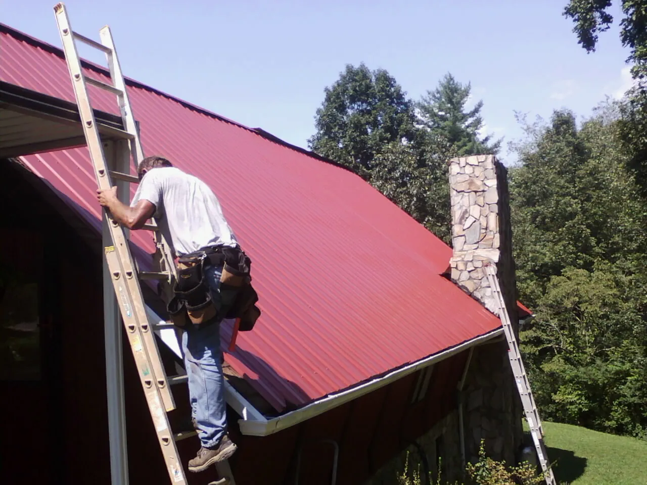 Roofer on a ladder beside a red metal roof and stone chimney — Soffit & Fascia Repair by Roofing Brothers in Blanchard